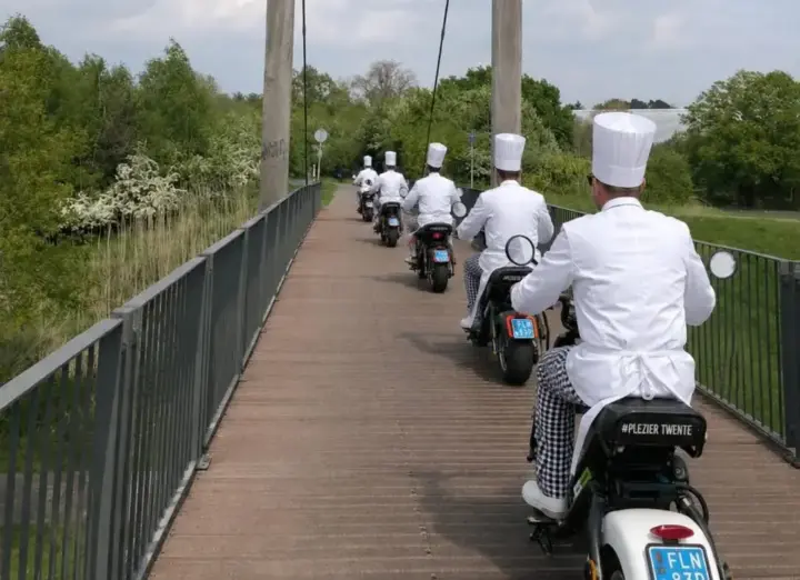 Koks met koksmutsen rijden op e-choppers over een hangbrug in het Twentse landschap bij Enschede