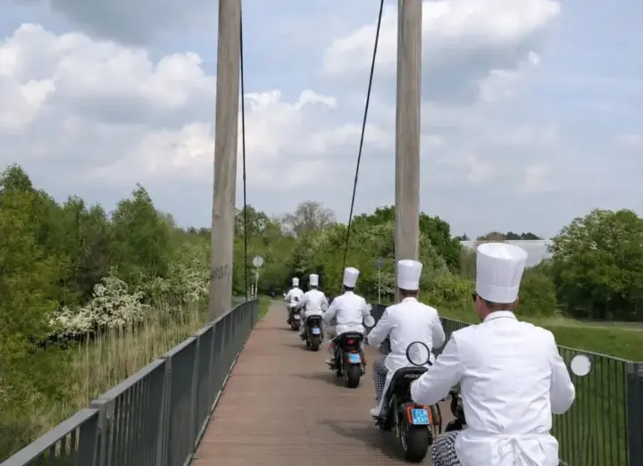 Collega's in koksoutfit rijden op e-choppers over een hangbrug in het Twentse landschap tijdens een personeelsuitje nabij Enschede